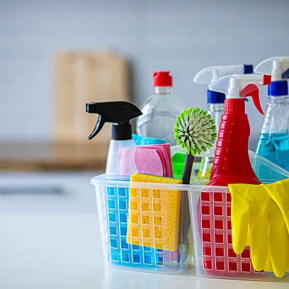 A plastic basket filled with various cleaning supplies including spray bottles, sponges, rubber gloves, and brushes placed on a white kitchen countertop. The background shows a modern kitchen with wooden and white cabinets, perfect for illustrating home cleaning, hygiene, or housekeeping concepts.