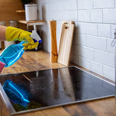A person wearing yellow rubber gloves is cleaning a black ceramic stove using a cleaning cloth. The image represents domestic chores, hygiene, and home cleanliness.